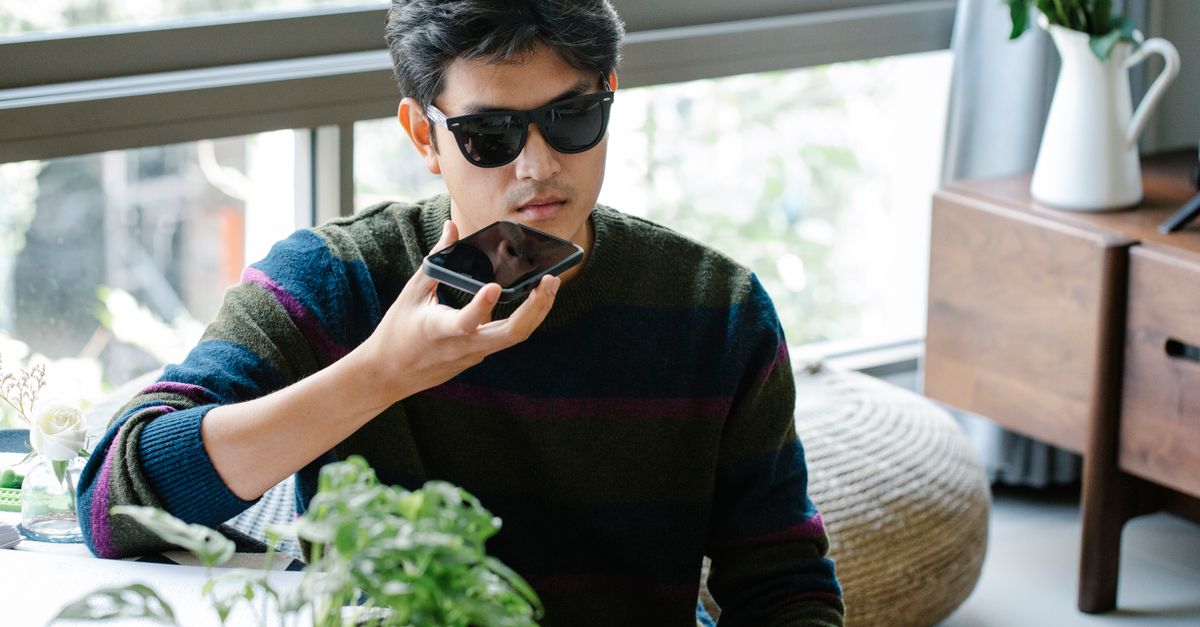 A man wearing sunglasses uses a smartphone's voice assistant in a cozy indoor setting.